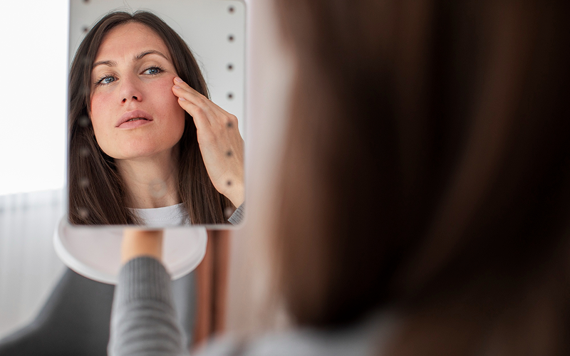 Woman examining facial skin redness in mirror.