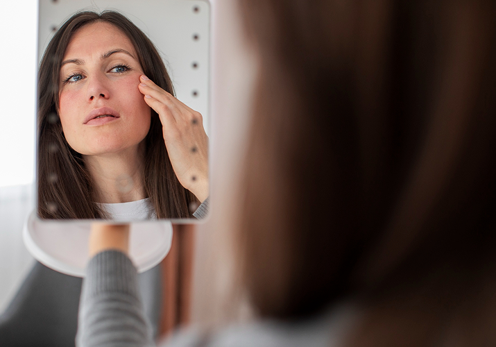 Woman examining facial skin redness in mirror.